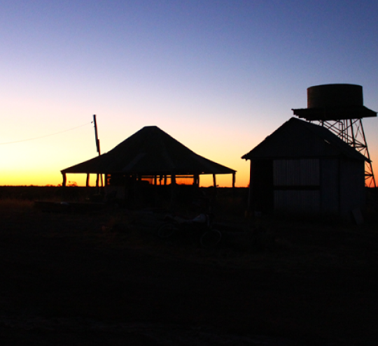 Farm homestead in sillouette at sunset or sunrise with water tank and windmill in the background.