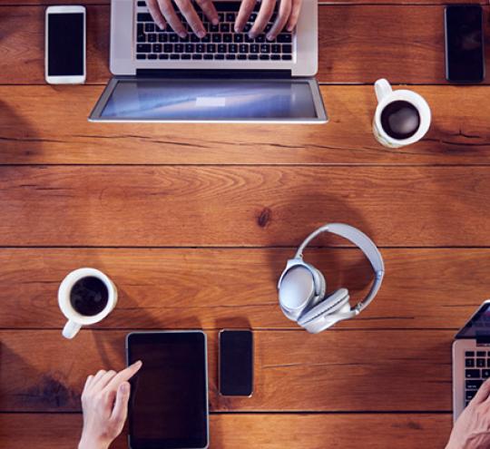 Image looking down on a table of people using laptops and mobile devices including three mugs of coffee and a pair of cordless headphones sitting in the middle of the table.