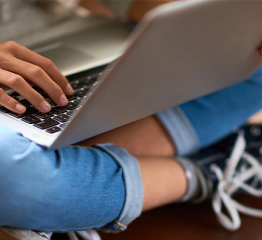 Young person wearing jeans and sneakers sitting cross-legged with a laptop