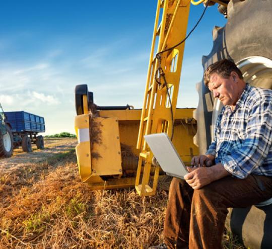Farmer counts yields on a laptop whilst sitting in the tyre of a tractor in the middle of a paddock with another blue tractor in the background.