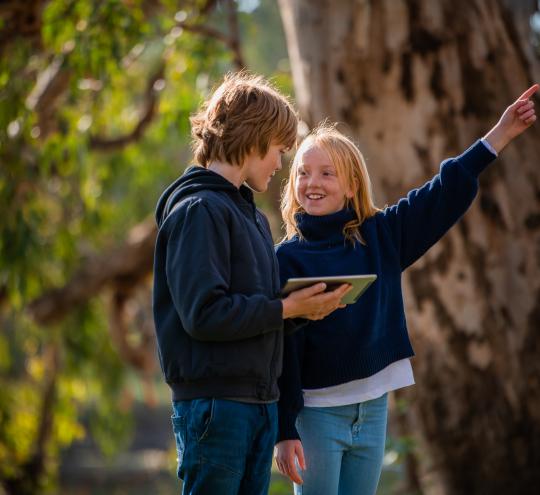 A young boy and girl standing outside in a park holding a digital tablet. 