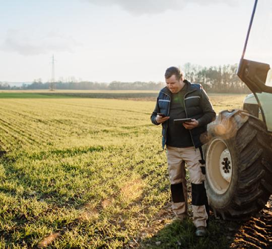 Man holding a mobile phone and tablet standing next to a tractor in a green field. 