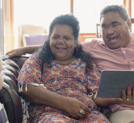 Man and woman sitting on a couch with a tablet looking at young grandson.