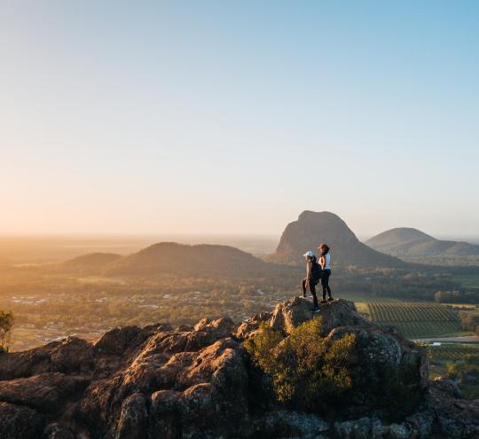 Two hikers stand on a hill-top looking into the distance. 