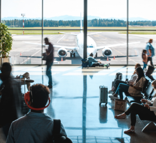 People waiting in a departures lounge of an airport terminal. A plane is visible outside large glass windows.