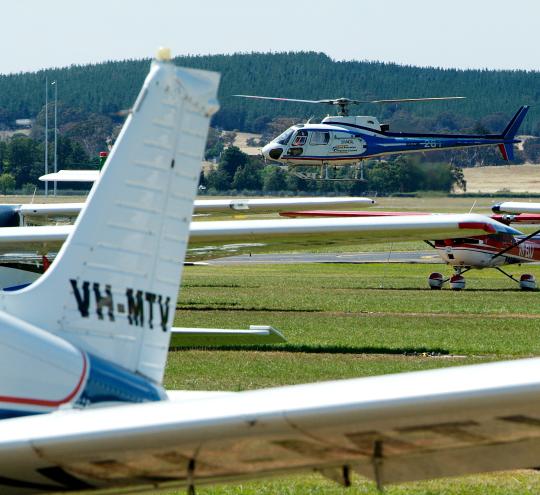 The wing and tail of a small aircraft at an airfield is in the foreground. In the background there is another small airplane and a hovering helicopter. 