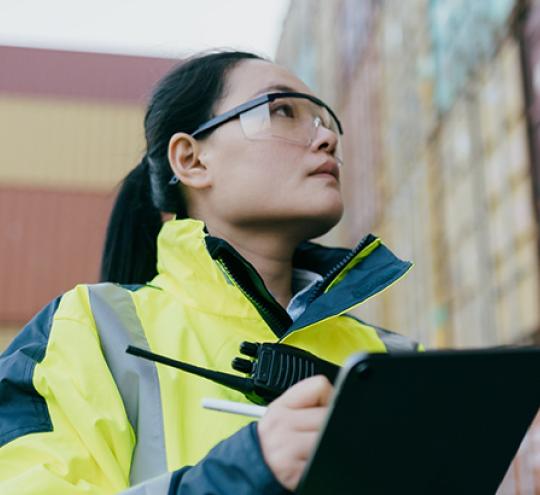 Asian female worker with clipboard and safety glasses looking up at a shipping containers stacked.