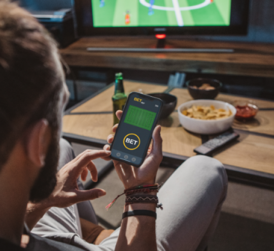 Man sitting on a couch watching a soccer match, holding a phone in his hand with a betting site open 