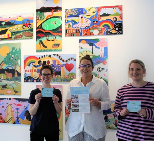 An image of three people involved with the Central Grampians Local Learning and Employment Network Inc’s ‘Ararat Schools Resilient Art Project’, to build positive educational outcomes and increase the resilience of young people in the community by delivering an art-based wellbeing program. The three people are smiling and standing in front of colourful artwork hung on a wall. Image credit: FRRR