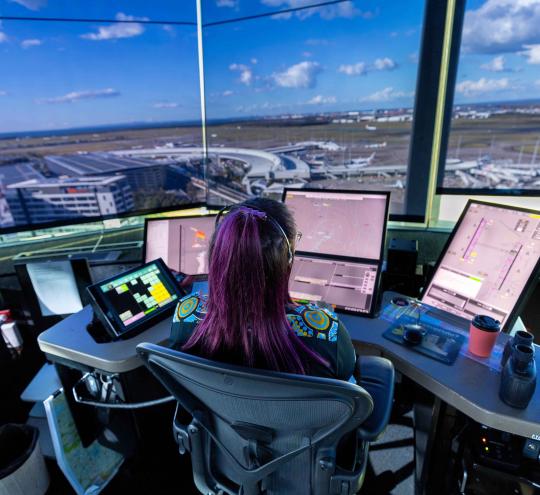 Photograph of an air traffic controller in a control tower managing multiple digital screens displaying radar, flight paths, and airport layout. Large windows provide a panoramic view of an airport terminal with several parked airplanes.