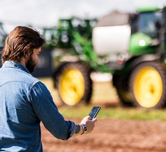 Farmer in a field looking at smartphone in front of a combine harvester.