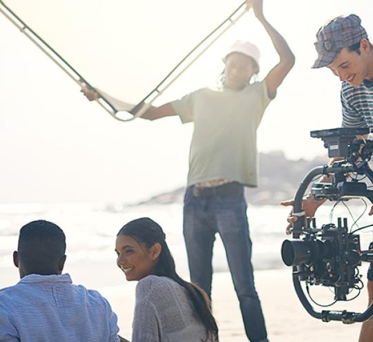 Screen crew filming couple sitting and talking at the beach.