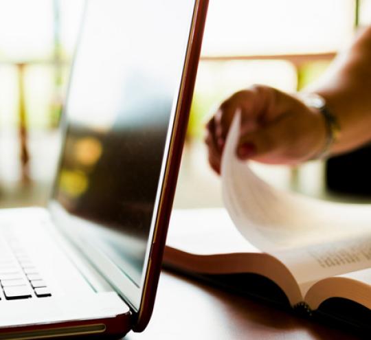 Pair of hands using a laptop on one side of a desk. On the other side of a desk is a hand flipping through a book.