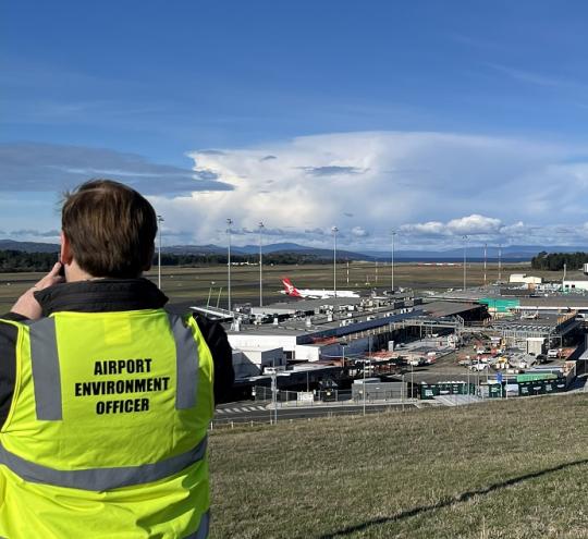 : image depicts the back of a man wearing hi vis that says, “Airport Environment Officer.” He looks out over an airport with terminal and hangar buildings visible in the distance, and an airplane parked on the runway behind the terminal.