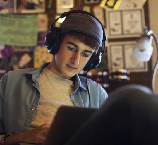 Young man wearing headphones and a backwards baseball cap working on laptop in his bedroom. 