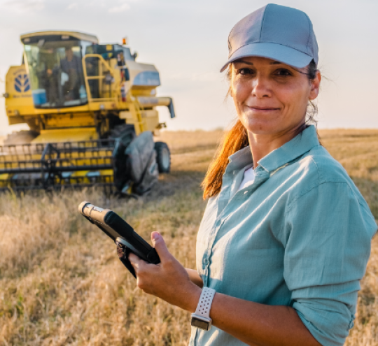 Female farmer holding a tablet in a field in front of a combine harvester. 