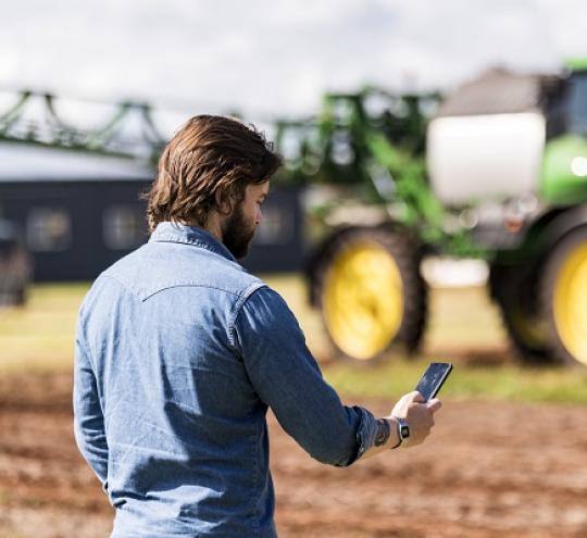 man standing in field using a mobile phone while observing a tractor