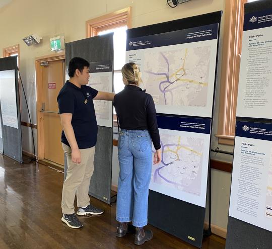 A man and woman look at a flight path map on display in a community hall