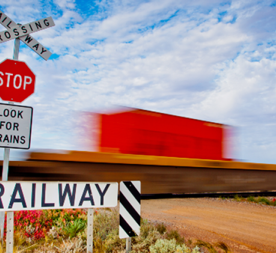 A stop and look for trains sign in the foreground with a blurry green, red and blue rail train in the background driving over a level crossing on a dirt road. 