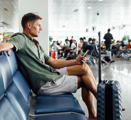 Image depicting man in shorts and collared shirt sitting in an airport terminal seating area, with carry-on baggage, looking at smartphone.