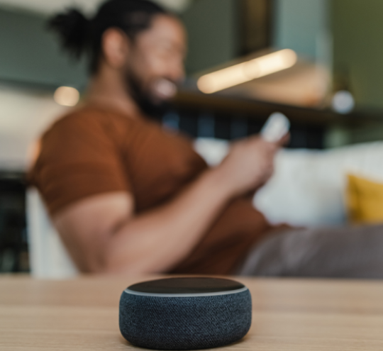 In the background a man sits on the couch, in the forefront a smartspeaker sits on a coffee table.