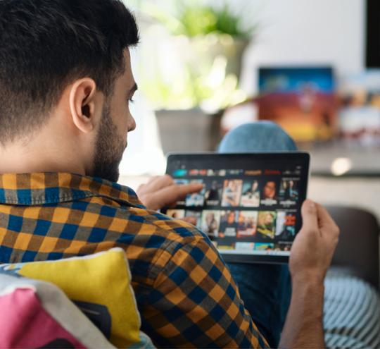 Man sitting on couch scrolls through titles available on a streaming service on an Ipad.