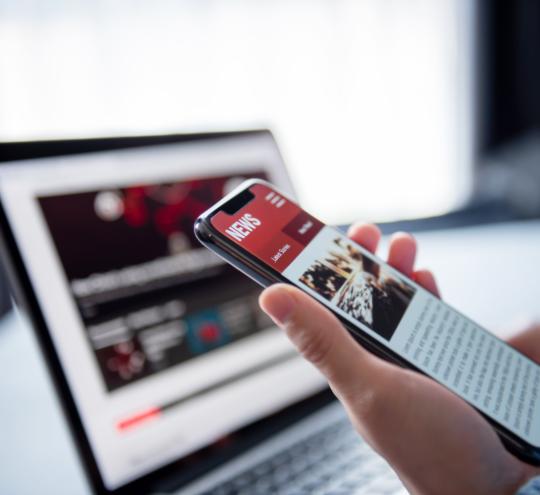 A person holding a mobile phone with a news website open, a laptop sits in the background with a website open.