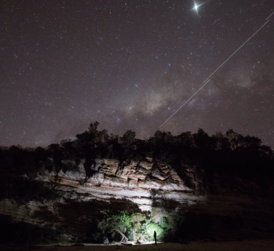 Campfire at cliffside looking up at a starry night and a low earth orbit satellite star train. 