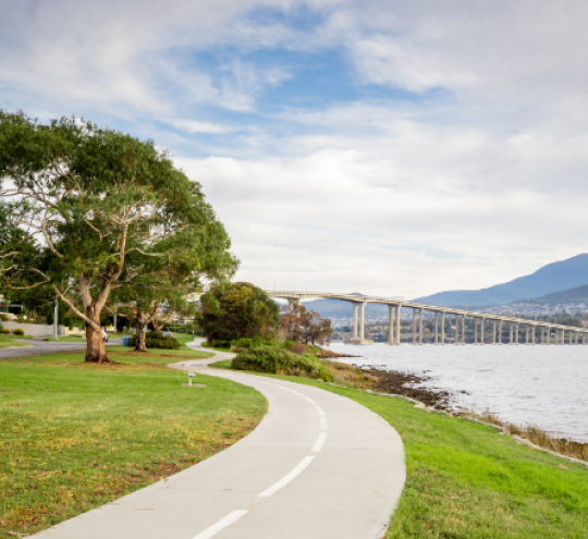 Shared bike and walking path, running alongside a road, hugging a body of water, with a bridge and mountains in the distance.