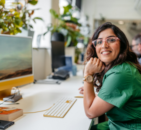 Woman seated at a desk in a modern office workspace, wearing glasses and a green outfit. A computer monitor, keyboard and indoor plants are visible, with a colleague and office area softly blurred in the background.