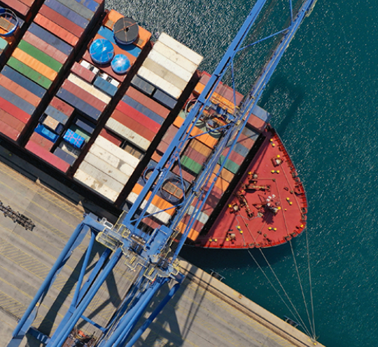 Looking down on a cargo ship with shipping containers, docked at the port next to gantry cranes. 
