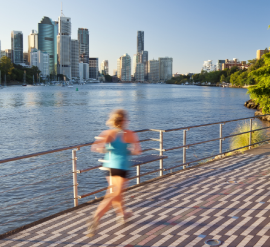 A woman jogs along a riverside footpath with colourful art sculptures in the foreground. In the background are established trees, and a city skyline across the river.