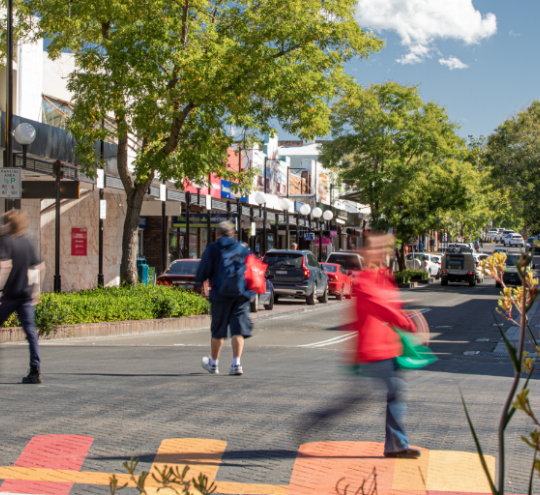 Pedestrians crossing regional city centre main street with community facilities and local businesses. 