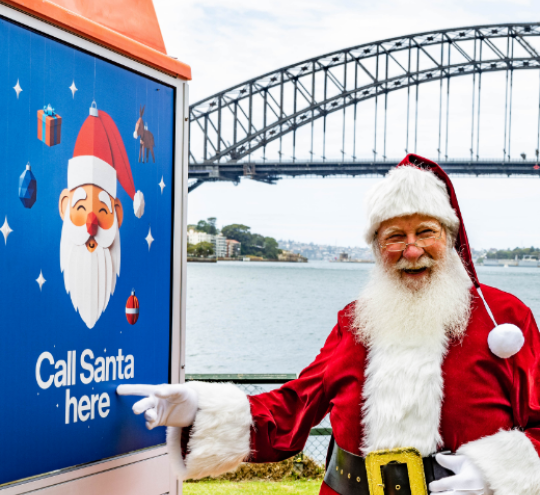 Santa pointing to a Telstra payphone with ‘Call Santa here’ printed on it in front of Sydney harbour.