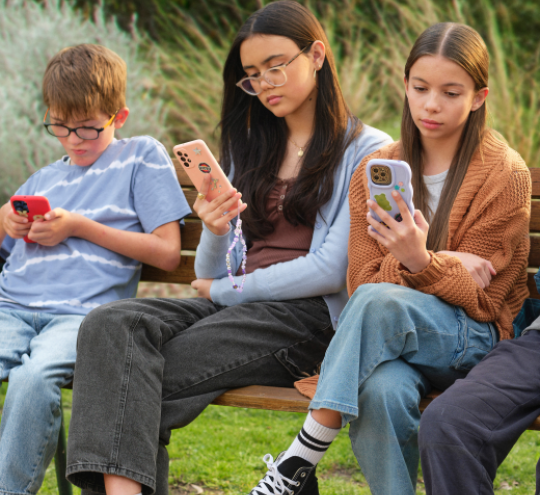 Three young people sitting on park bench, looking at their phones