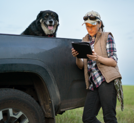 Alt text for image Short visual description of image for screen readers used by people who are vision impaired	Young female farmer, leaning against her car in a field while looking at her tablet. Dog sitting in the back of the truck watching.