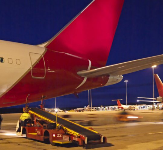 The rear wing of a large passenger airplane docked at an airport in the evening, with ground handling vehicle in the foreground and other planes in the background.