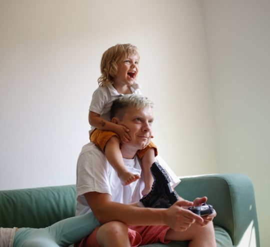 Father sitting on a couch playing video games with child sitting on his shoulders