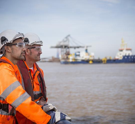 Two maritime workers looking out to sea with a ship in the background.