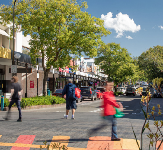 Four people walking on a street in a regional town setting, featuring older-style buildings with store-fronts, cars parked out the front of the stores, and trees along the sides of the street. 