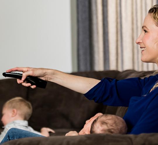 Woman sitting on a couch with two toddlers with a remote in her hand. She is smiling.