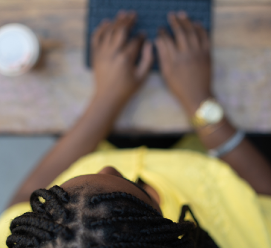 Top down view of person with black braided hair, wearing a yellow shirt typing on a laptop. There is a takeaway hot drink cup to their left.