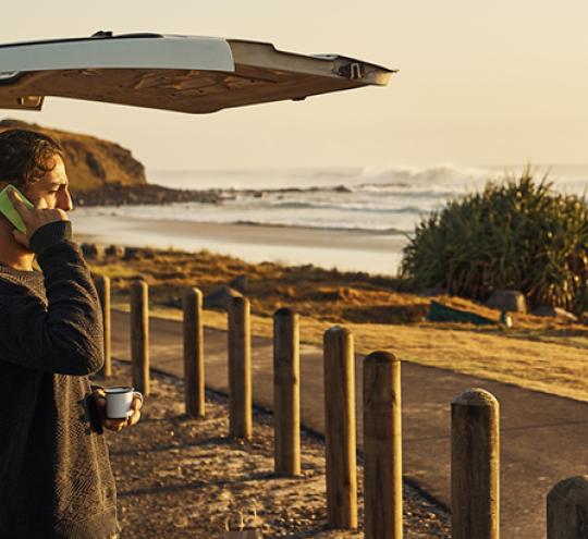 Man standing at the end of a van looking over a beach and talking on a mobile phone.