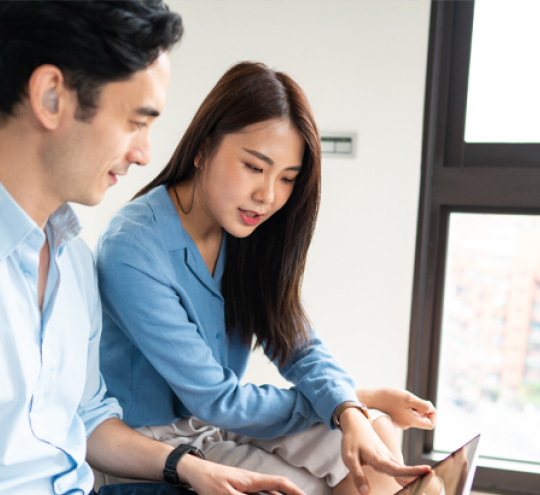 Young man wearing hearing aid working on a laptop that is open on his lap next to a young woman who is looking at the laptop and holding a phone.