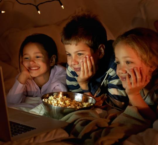 Three children in a tent with a bowl of popcorn watching a laptop.