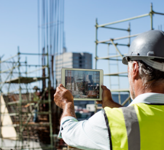 A worker in high vis jacket and hard hat holds up a tablet in front of them looking at at construction site
