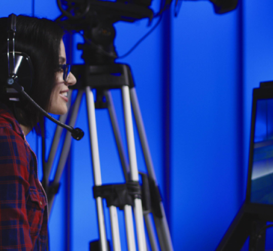 Film set with female director and cameraman looking at TV screen which has two people sitting on chairs talking.