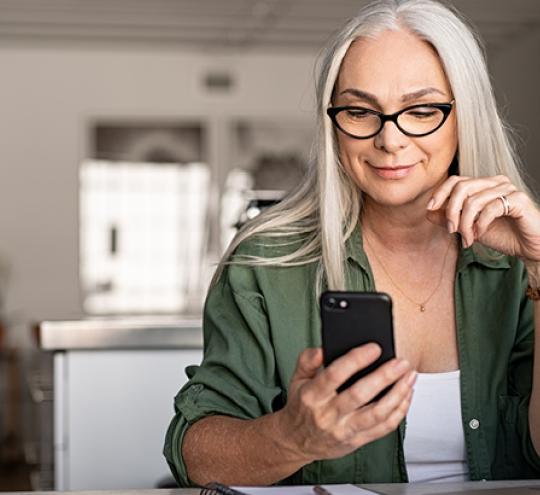 Woman sitting in front of laptop and looking at a mobile device in her hand.
