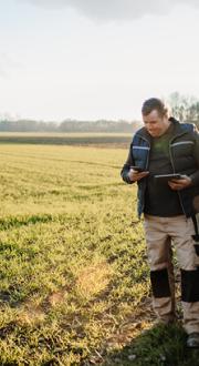 Man holding a mobile phone and tablet standing next to a tractor in a green field. 