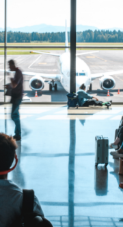 People waiting in a departures lounge of an airport terminal. A plane is visible outside large glass windows.